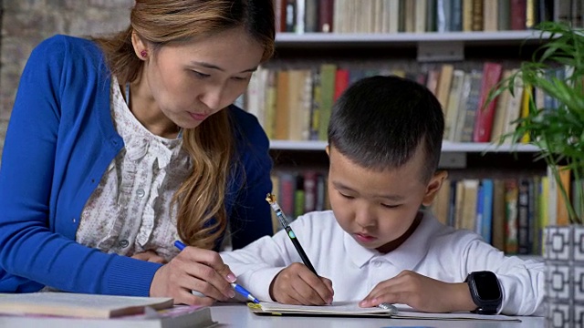 亚洲小男孩做作业与年轻的母亲，妇女帮助孩子，坐在桌子后面，书架的背景视频购买