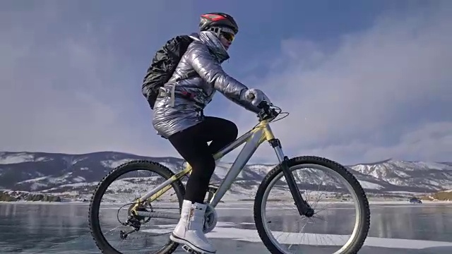 一个女人在冰上骑自行车。这个女孩穿着银色的羽绒服，背着自行车背包，戴着头盔。结冰的贝加尔湖的冰。自行车的轮胎上覆盖着特殊的尖钉。视频素材