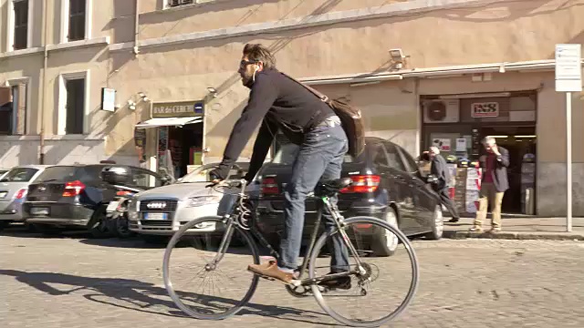 在阳光明媚的日子里，一名年轻的潮男骑着自行车在罗马市中心的历史建筑旁行走，他还戴着墨镜视频素材