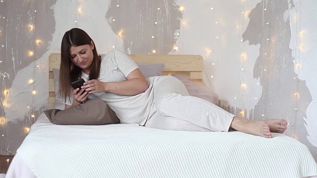 快乐美丽的女孩在睡衣和长发享受手机躺在家里的床上视频素材