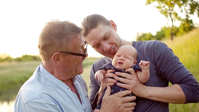 爷爷和爸爸充满爱意地看着这个月大的婴儿。刚出生的婴儿和他的祖父和父亲在河边度假。日落视频素材