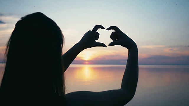 美丽的女人在日落时用手做心形的女孩用橙色的太阳耀斑举着爱的象征手势视频素材
