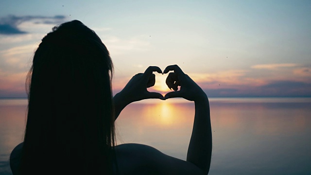 美丽的女人在日落时用手做心形的女孩用橙色的太阳耀斑举着爱的象征手势视频素材