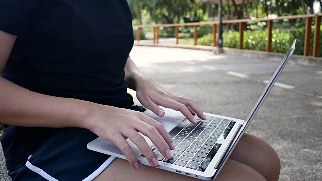 美丽的亚洲年轻女子坐在公园的长椅上使用笔记本电脑。迷人的女性看她的电脑在温暖的春天的一天。女性生活方式的概念。视频下载