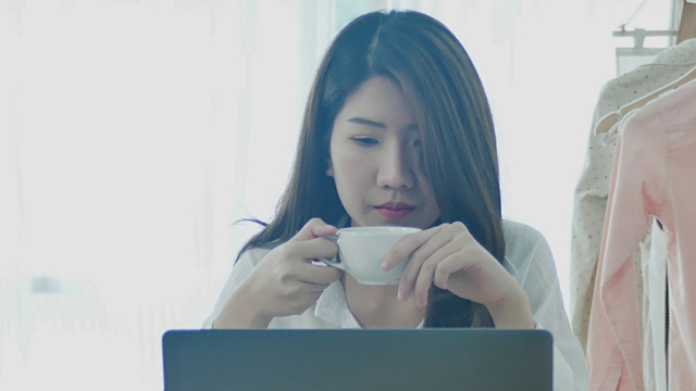 美丽的年轻微笑的女人工作在笔记本电脑，同时享受喝温暖的咖啡坐在家里的客厅。享受在家的时光。在家庭办公室工作的亚洲女商人。视频下载