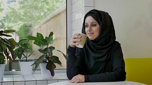 年轻美丽的穆斯林妇女戴着头巾喝着咖啡，微笑着坐在咖啡馆里视频下载