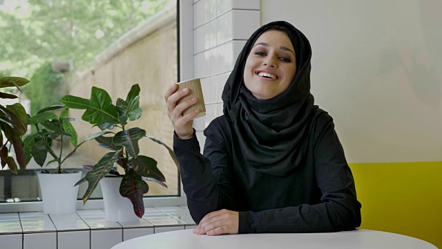 年轻迷人的穆斯林妇女戴着头巾喝着咖啡，看着镜头，笑着坐在咖啡馆里视频下载