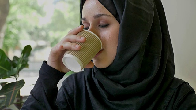 年轻的穆斯林妇女戴着头巾坐在咖啡馆里，喝着咖啡，微笑着看着镜头视频下载