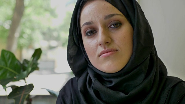 这是一幅年轻的穆斯林妇女戴着头巾坐在咖啡馆里，直视着镜头，沉思着，忧心忡忡视频下载