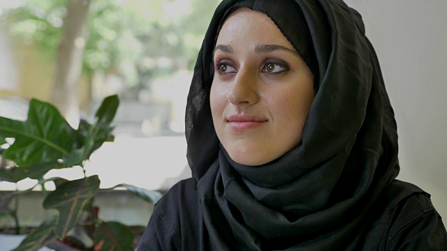 年轻美丽的穆斯林妇女戴着头巾坐在咖啡馆里，向前看，然后在镜头里微笑视频下载