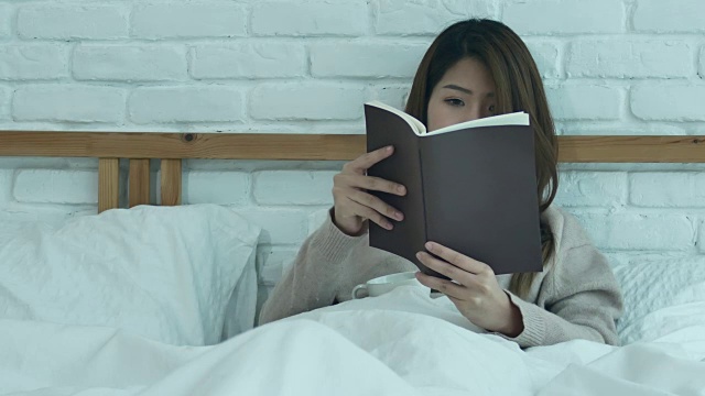 美丽的亚洲女人享受喝温暖的咖啡和看书在床上在她的卧室。亚洲女性穿着舒适的毛衣捧着一本书和一杯咖啡。生活方式亚洲女性在家的概念视频下载