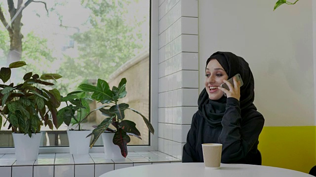 年轻漂亮的穆斯林妇女戴着头巾在打电话，微笑着，坐在咖啡厅里，心情愉快视频下载
