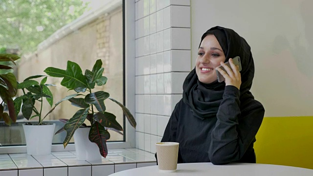 年轻迷人的穆斯林妇女戴着头巾，一边打电话一边大笑，坐在咖啡馆里，很开心视频下载