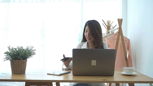 慢动作-美丽的年轻微笑的亚洲女人在笔记本电脑上工作，而坐在家里的客厅。亚洲商业女性在她的家庭办公室使用电话工作。享受在家的时光。视频下载