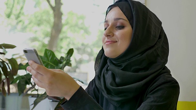 年轻漂亮的穆斯林妇女戴着头巾坐在咖啡厅里，拿着电话，用手机聊天，面带微笑视频下载
