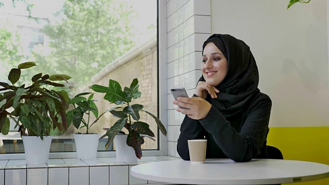 年轻美丽的穆斯林妇女戴着头巾，拿着电话微笑着，坐在咖啡厅里，心情愉快视频下载