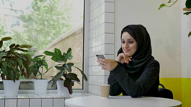 年轻漂亮的穆斯林妇女戴着头巾拿着电话，用手机聊天，微笑着，坐在咖啡馆里，开心视频下载