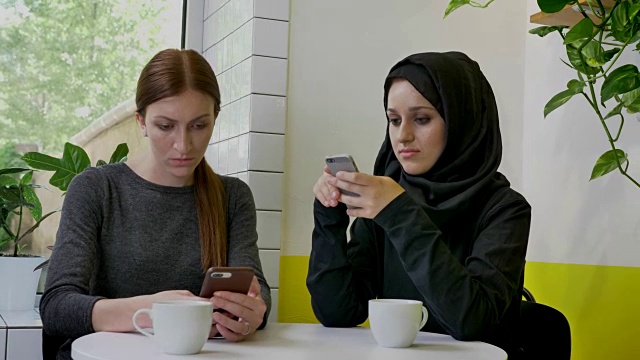 两个年轻漂亮的女人坐在咖啡馆里打着电话，其中一个戴着头巾，坚定而专注视频下载