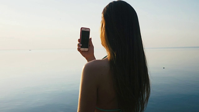 漂亮的年轻女子与她的智能手机在日落海滩附近的海边度假。慢动作视频下载