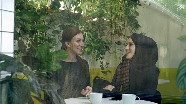 两名年轻女子坐在咖啡馆里，其中一名是戴着头巾的穆斯林妇女，在外面看着她们有说有笑视频下载