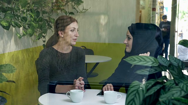 两个年轻美丽的女人坐在咖啡馆，其中一个是穆斯林妇女戴着头巾，有说有笑，从街上看视频下载