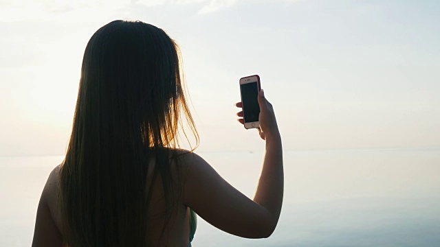 漂亮的年轻女子与她的智能手机在日落海滩附近的海边度假。慢动作视频下载
