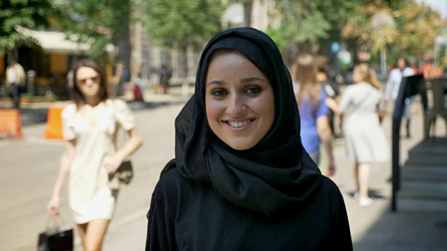 年轻美丽的穆斯林妇女戴着头巾站在城市街道上，微笑着看着镜头视频下载