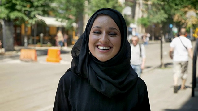 年轻迷人的穆斯林妇女戴着头巾站在城市街道上，笑着看着镜头视频下载