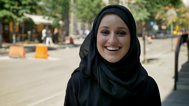 年轻漂亮的穆斯林妇女戴着头巾站在城市街道上，直视镜头，笑着视频下载