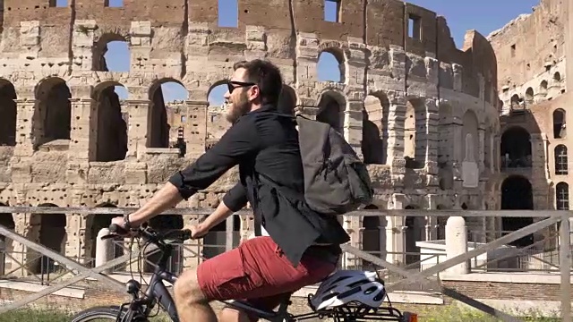 三个年轻的朋友游客在阳光明媚的日子里骑着自行车在罗马市中心的罗马斗牛场周围的道路上慢镜头摄影机汽车稳定摄影机视频素材