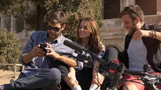 在阳光明媚的日子里，三个快乐的年轻朋友骑着自行车和背包，坐在罗马斗牛场用智能手机自拍视频素材