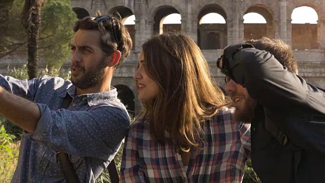 三个快乐的年轻朋友，游客在罗马斗牛场用智能手机拍有趣的自拍照，在日落时用树木和慢动作摄影机在山上做鬼脸视频下载