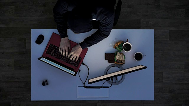 一个穿着黑帽衫的年轻人在晚上侵入笔记本电脑，坐在桌子后面拿着笔记本电脑和显示器，上图视频素材