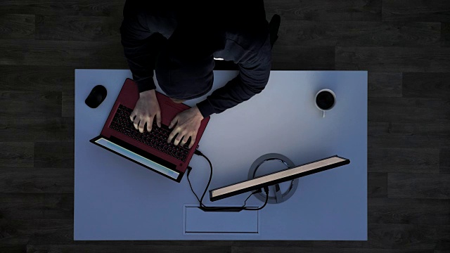 一名穿着黑色帽衫的年轻人坐在一张白色的桌子后面，伸伸手指在笔记本电脑上打字，看着显示器视频素材