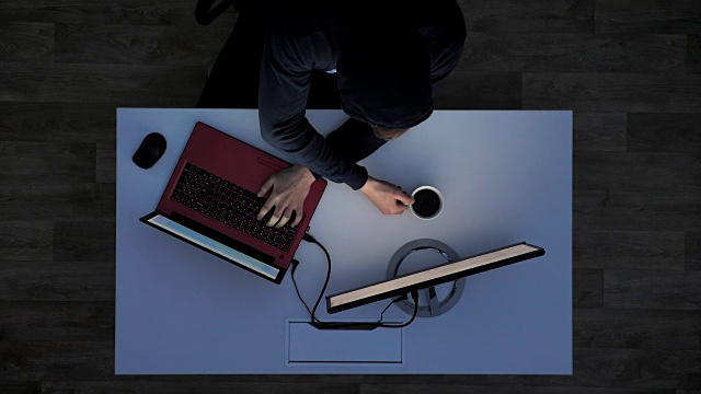 一个穿着黑色帽衫的年轻人坐在白色的桌子后面，喝着咖啡，在笔记本电脑上打字，上图视频素材