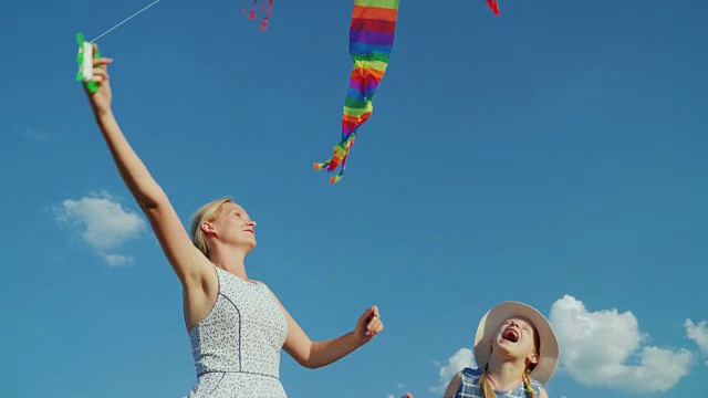 一个无忧无虑的女人和她的女儿玩，一个女孩试图抓住一个空中风筝。以纯净的蓝天为背景视频素材