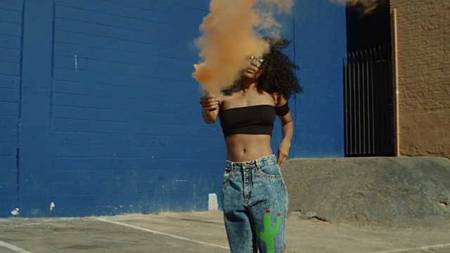美丽的年轻女子拿着五颜六色的烟雾弹在蓝色的墙壁上跳舞视频素材
