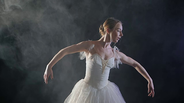 (中景)一个美丽的年轻芭蕾舞演员在聚光灯下优雅地旋转。她穿着白色轻盈的现代芭蕾舞裙。性感的女人在冒烟的舞台上。慢动作视频素材