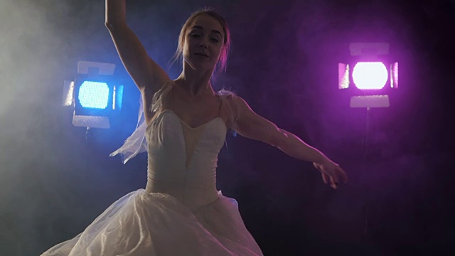 性感芭蕾舞女演员身穿古典或现代芭蕾舞元素的白色芭蕾舞裙，在黑暗中旋转，泛光灯背景下有蓝色和紫色的光和烟雾视频素材