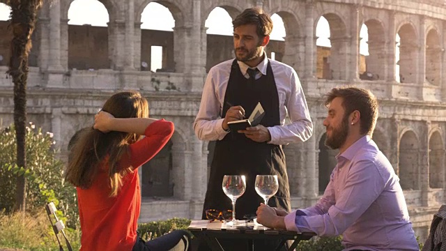 日落时分，一对浪漫优雅的情侣坐在罗马斗牛场前的餐厅桌前，和服务生一起点餐和饮料视频下载