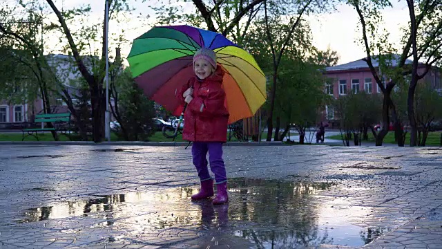可爱的小女孩拿着五颜六色的雨伞在夕阳下的公园里的水坑里跳跃。视频下载