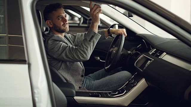 一个严肃的年轻人正坐在汽车陈列室的新车里，按下控制面板上触摸汽车部件的按钮检查电子设备。购买汽车的概念。视频下载