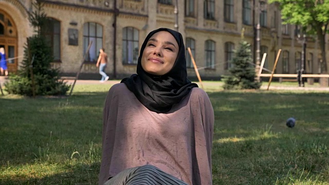 年轻快乐的穆斯林女孩戴着头巾坐在阳光下的草坪上，看着镜头，建筑在背景上，宗教观念，放松观念视频下载
