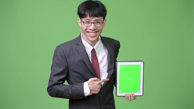 年轻快乐的亚洲商人向相机展示数码平板电脑，并指着手指视频下载