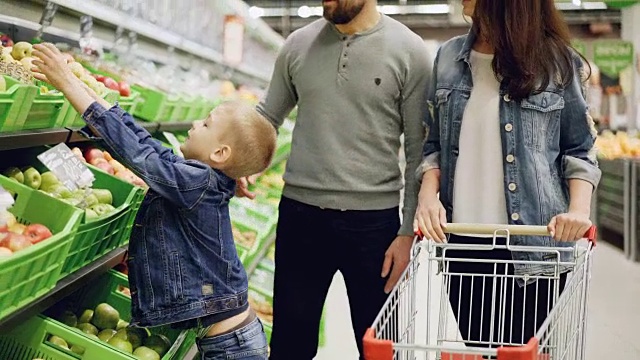 快乐的年轻家庭母亲，父亲和孩子在超市买水果，把菠萝放在购物车里，交谈和微笑。食品商店和人的概念。视频下载