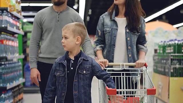 快乐的年轻家庭妈妈、爸爸和小男孩一起走在超市里，推着购物车四处张望，指着食物说着话。视频下载