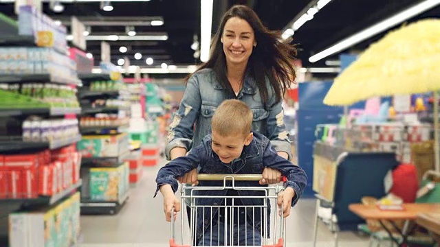 快乐慈爱的妈妈和她可爱的儿子在超市里玩得很开心，她推着购物车跑着，小男孩站在上面，人们都在笑。视频下载