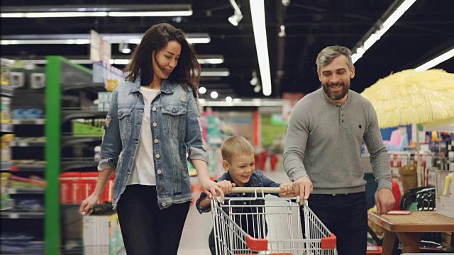 慢镜头，爸爸、妈妈和孩子推着购物车跑过超市，笑着。乐趣在商店和人的概念。视频下载
