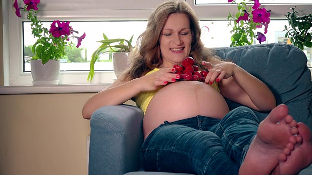 坐在沙发上微笑着吃草莓的美丽孕妇视频素材