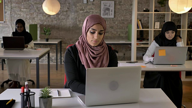 三个年轻的穆斯林妇女戴着头巾坐在现代化的办公室里工作，穆斯林女性工作者看着镜头微笑着视频素材
